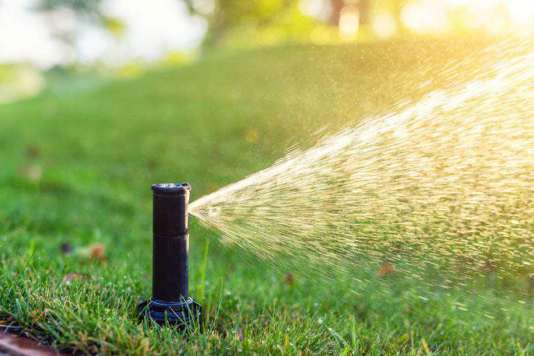 Close-up automatic garden watering system with different sprinklers installed under turf. Landscape design with lawn hills and fruit garden irrigated with smart autonomous sprayers at sunset time.