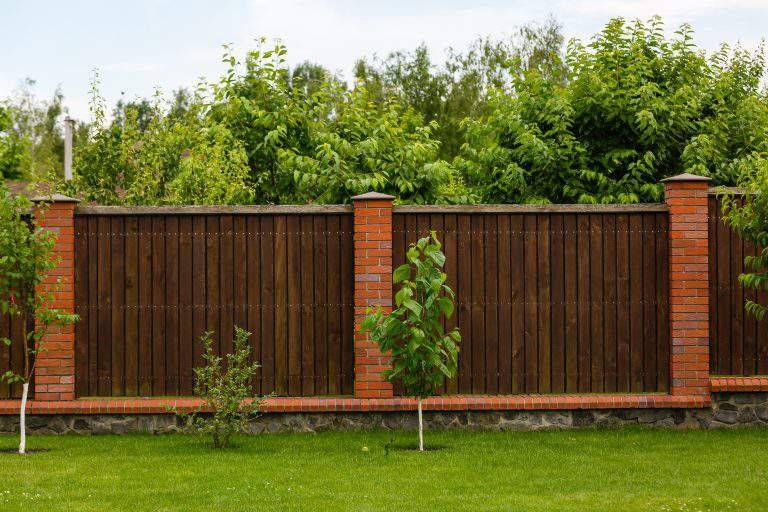 New wooden fence with massive stone brick pillars. Green lawn and trees, daytime